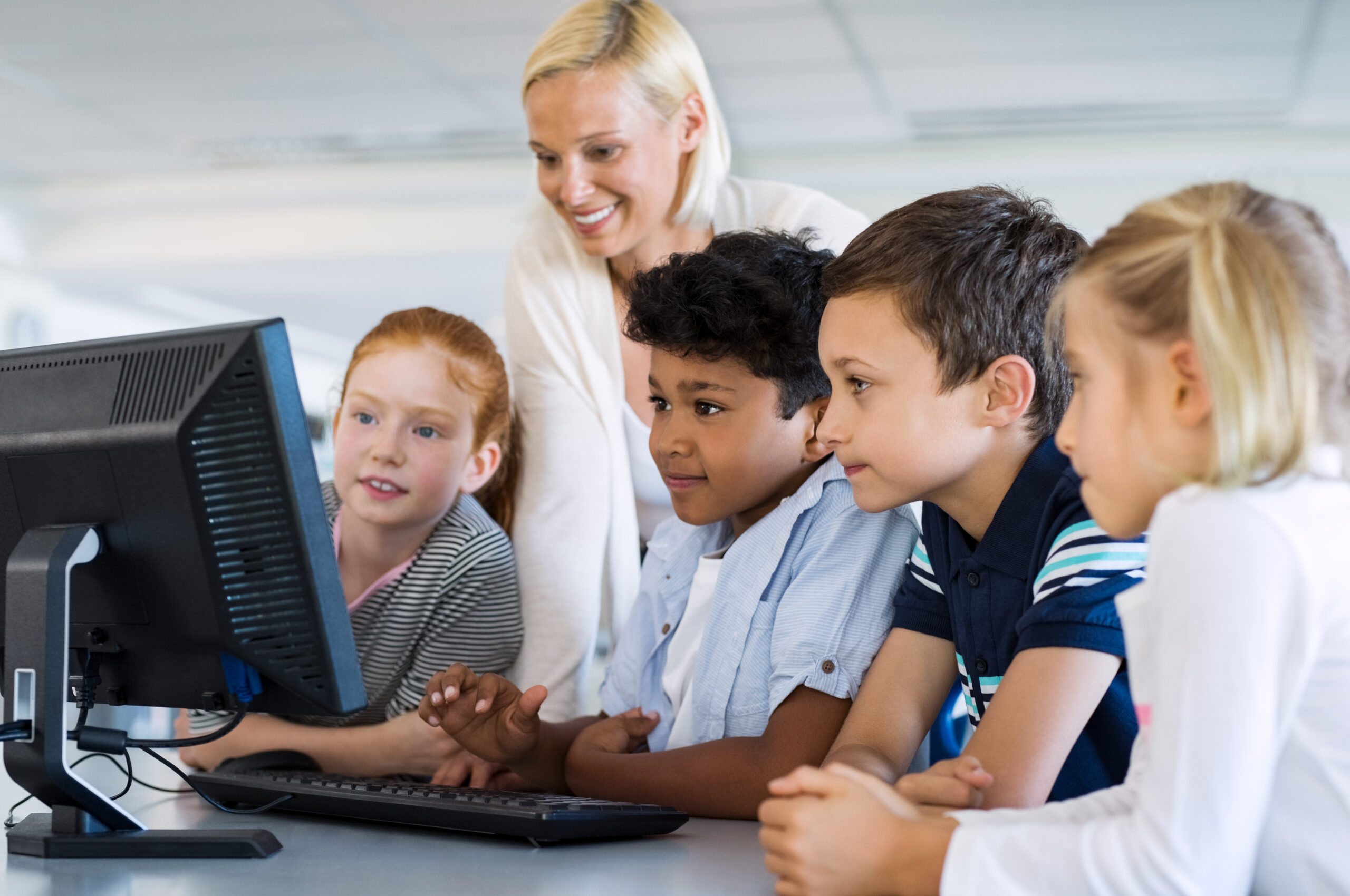 Teacher,Helping,Multiethnic,Children,At,Computer,Terminal,In,Primary,School.